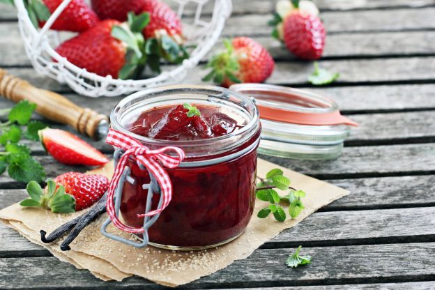 strawberry jam in a glass jar with strawberries around it on a wooden picnic table