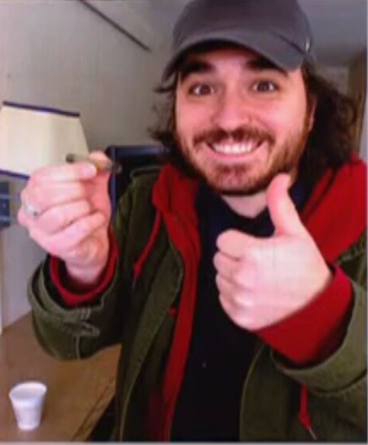 a man with long hair wearing a baseball cap and giving the thumbs up while sitting at a table