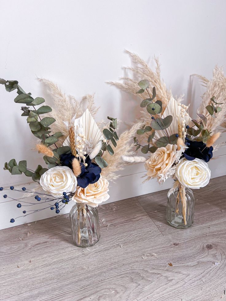 two vases filled with flowers and greenery on top of a wooden floor next to a wall