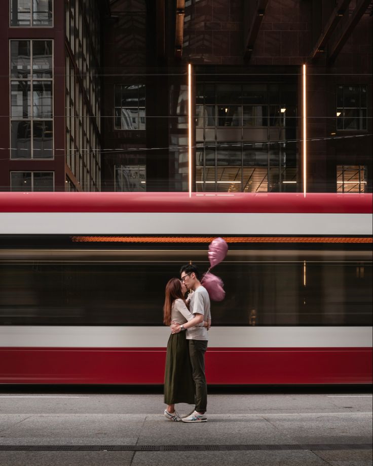 a man and woman standing next to each other in front of a train with pink balloons