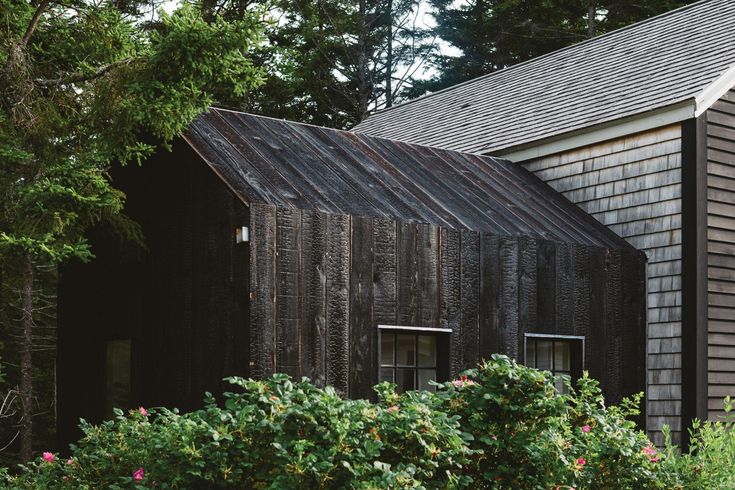 an old wooden building in the middle of some trees and bushes with flowers around it