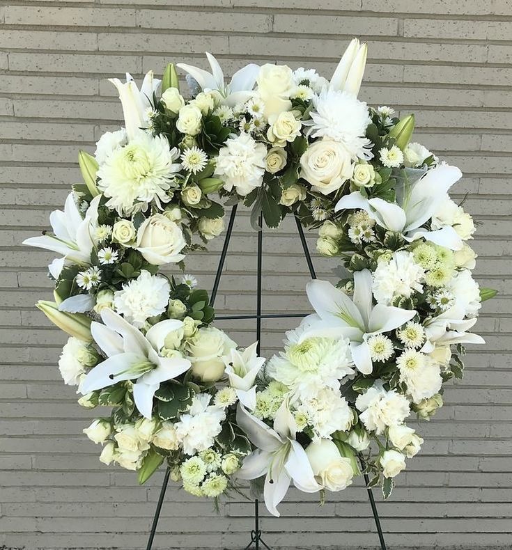 a wreath with white flowers and greenery in front of a gray brick wall on a metal stand