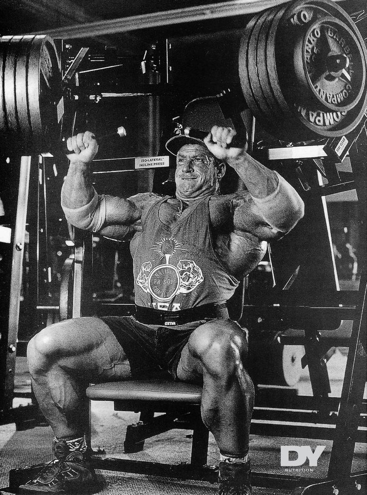 a man squats on a bench in front of a barbell rack and raises his arms