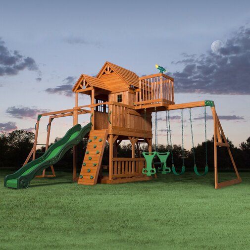 a wooden playset with a green slide and swing set in the grass at dusk