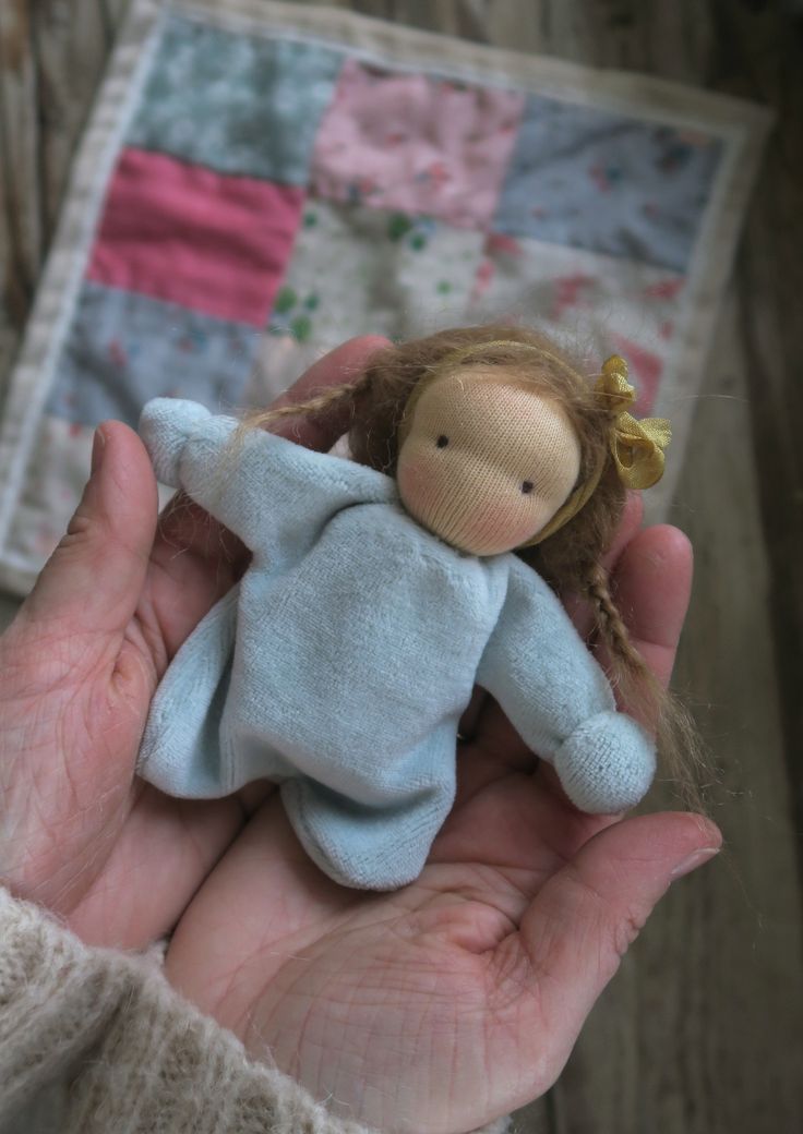 a hand holding a small doll in it's left hand on top of a quilt