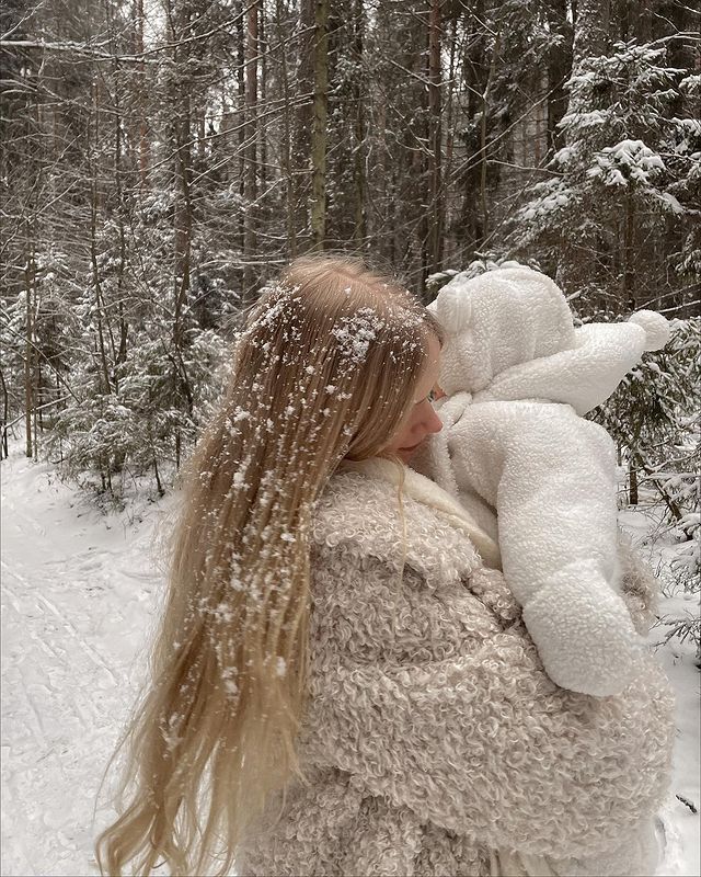 a woman is holding a stuffed animal in the snowy woods with snow falling all around her