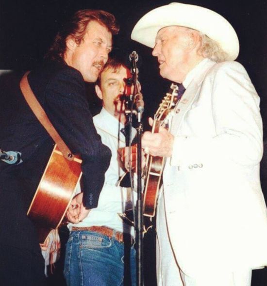 two men standing next to each other holding guitars and singing into microphones while another man plays guitar behind them