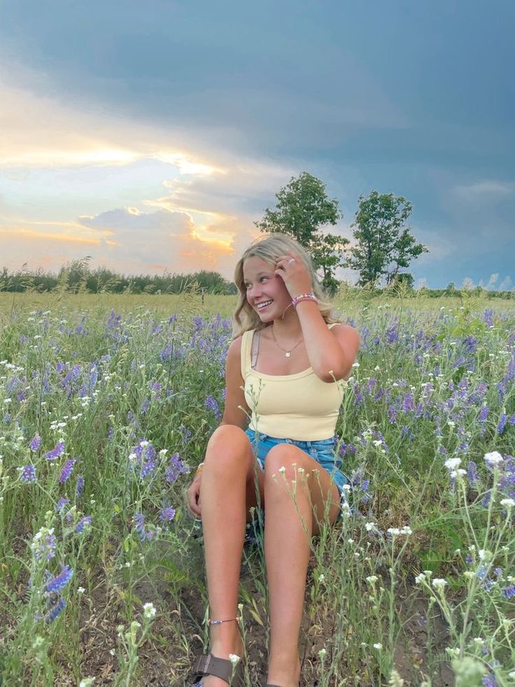 a woman sitting in the middle of a field with flowers on her knees and one hand up to her head