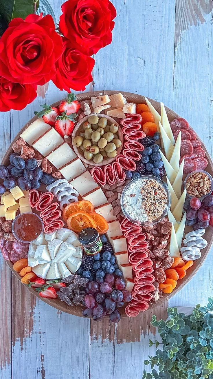 a platter filled with cheese, crackers, fruit and nuts next to red roses