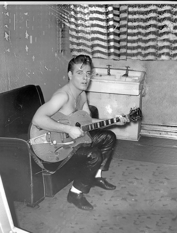 a man sitting in a chair holding a guitar and looking at the camera with an open suitcase behind him