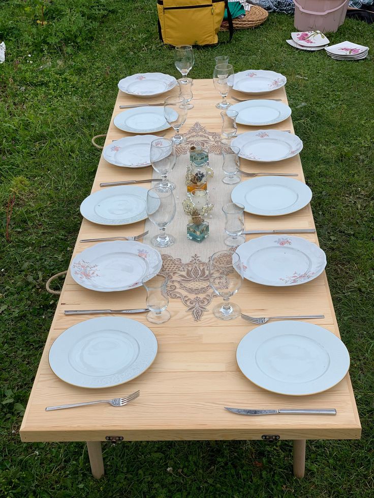 a long table set up with white plates and silverware on it in the grass