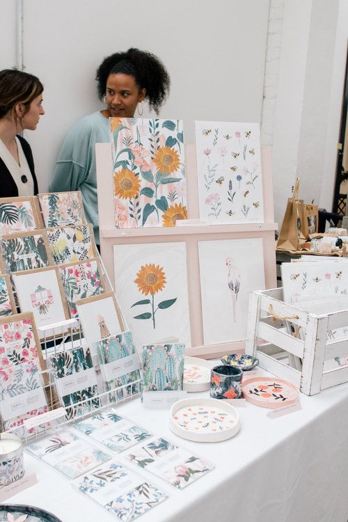 two women standing next to a table full of cards and artwork on it's sides