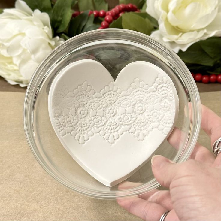 a person holding a heart shaped dish in front of some white flowers and greenery