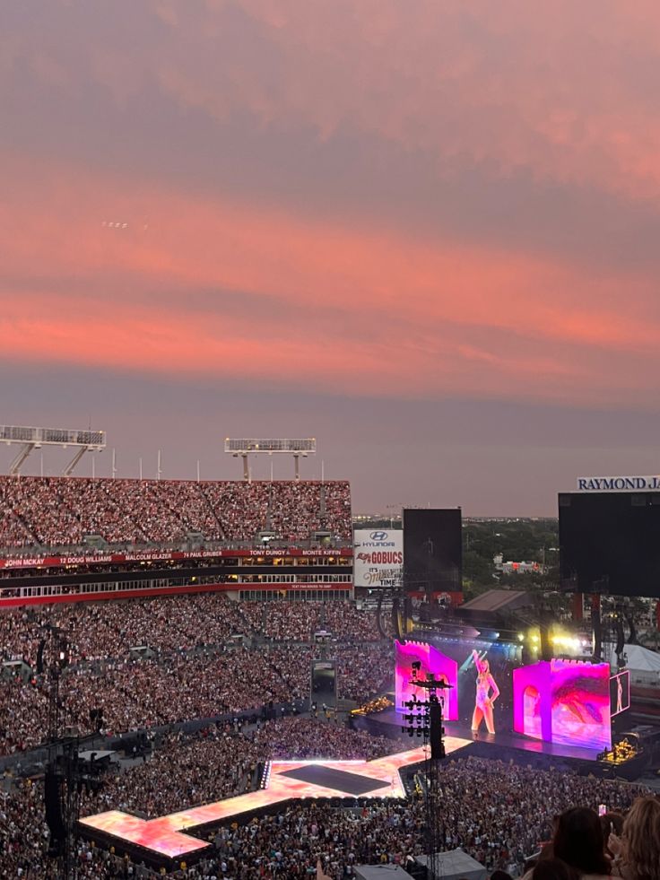 a large crowd is watching a concert on stage in front of a pink and purple sky