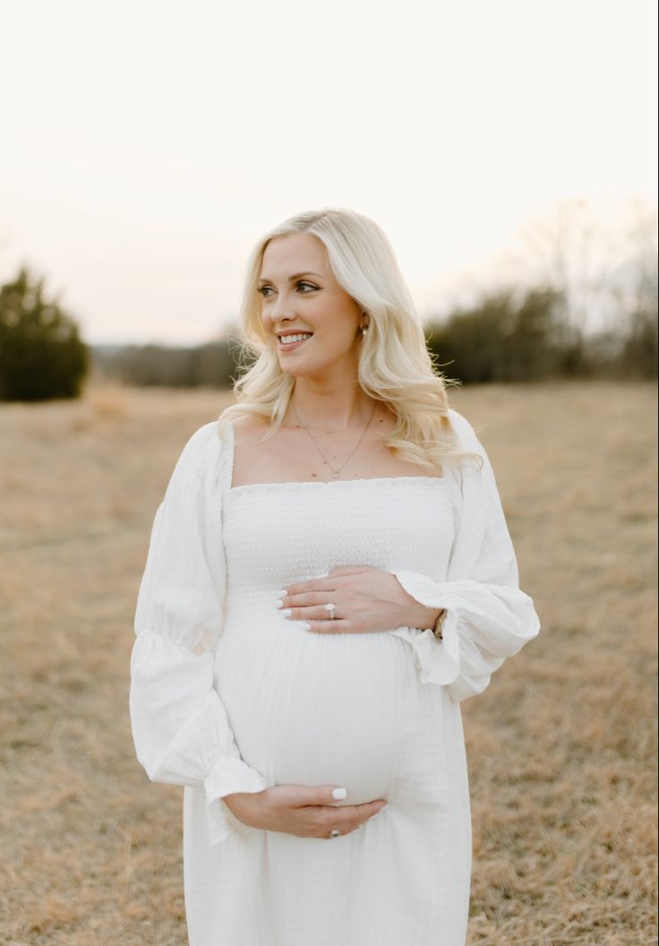 a pregnant woman in a white dress standing in a field with her hands on her belly