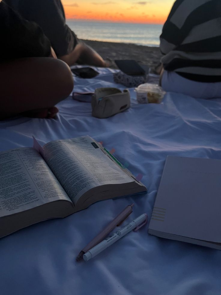 two people are sitting on the beach with an open book and pen in front of them