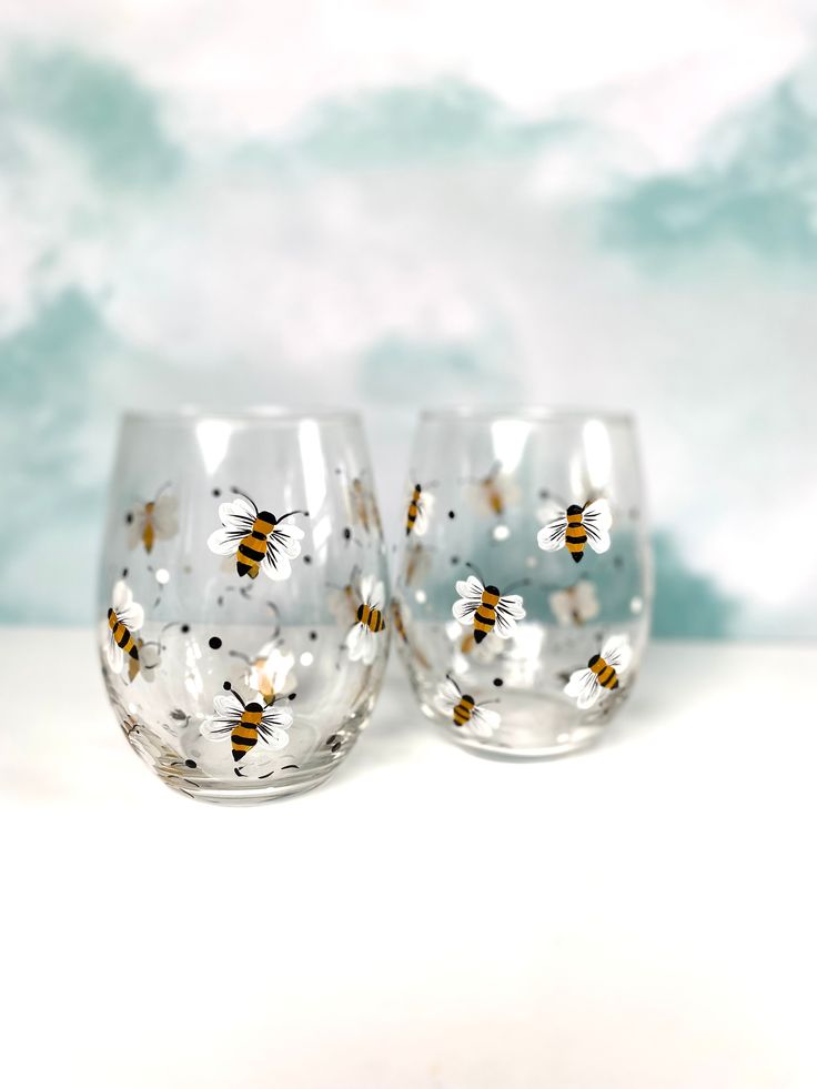 two glasses with bees painted on them sitting next to each other in front of a blue and white background