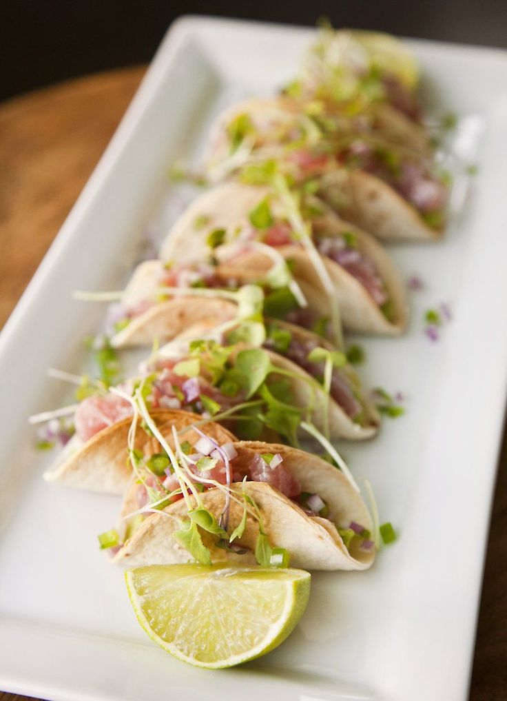 several tacos are lined up on a white plate with lime wedges and garnishes