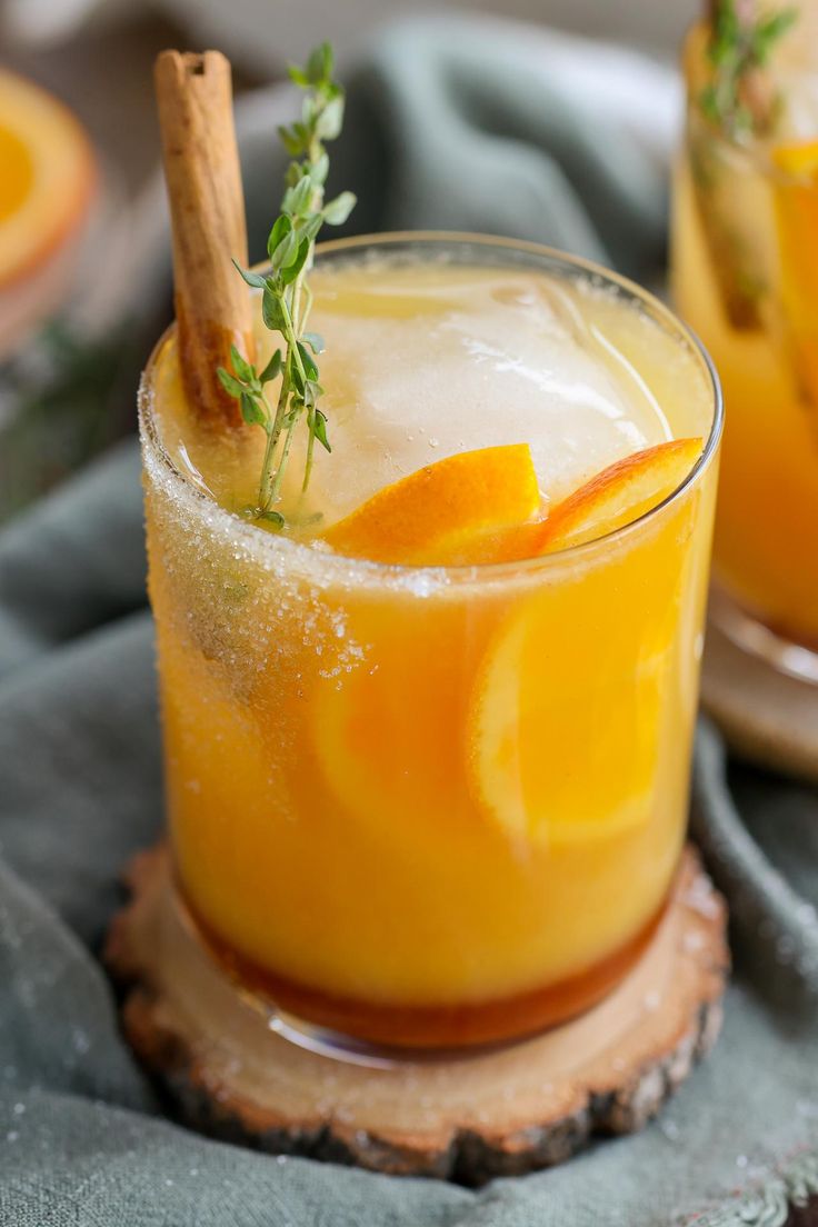 two glasses filled with orange juice and garnished with fresh herbs, sitting on a wood slice