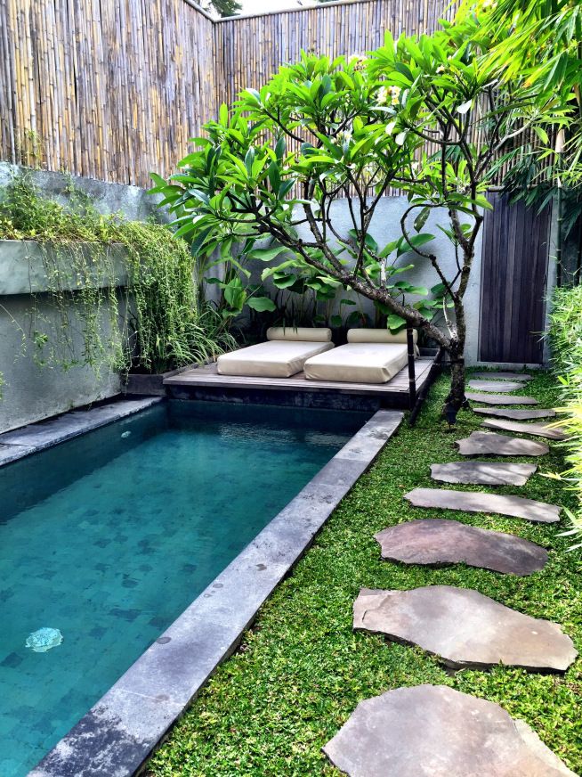 a small backyard with a swimming pool surrounded by greenery and stone stepping stones in the grass