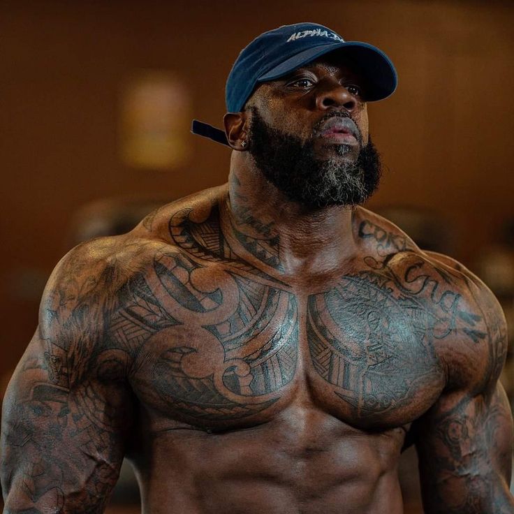 a shirtless man with tattoos on his chest and beard wearing a baseball cap is looking off to the side