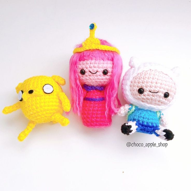 three crocheted dolls sitting next to each other on a white surface, one with pink hair and the other with blue eyes