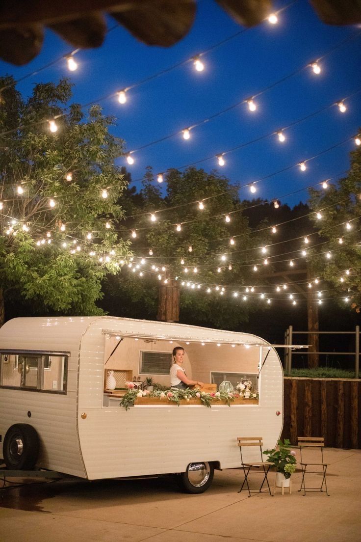 a white trailer with lights hanging from it's sides and a woman cooking outside