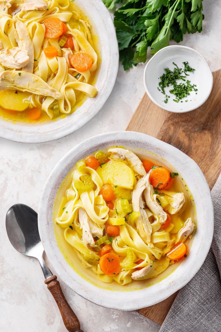 two bowls filled with chicken noodle soup on top of a wooden cutting board