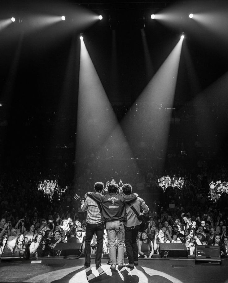 the backs of three men standing on stage with their arms around each other in front of an audience