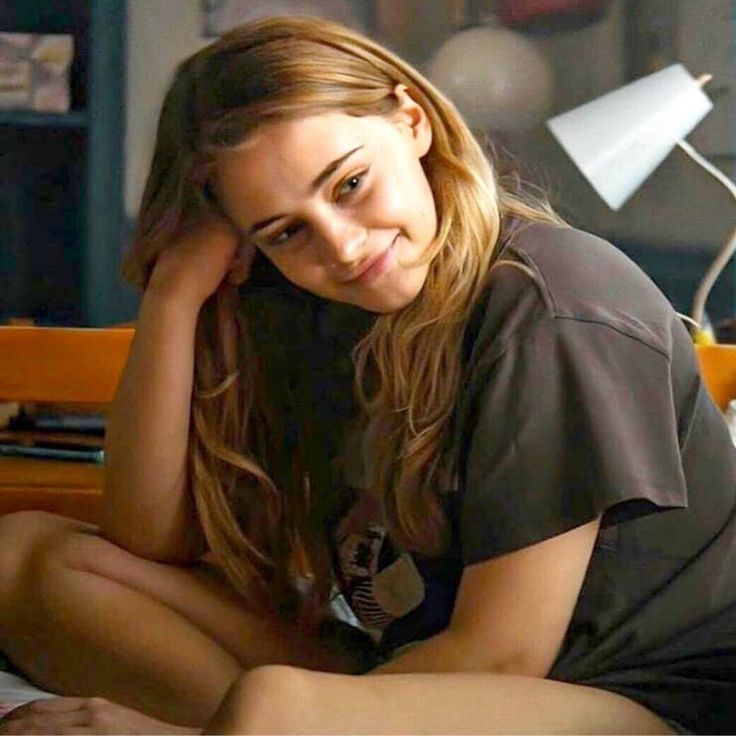 a young woman sitting on top of a bed next to a desk with a lamp