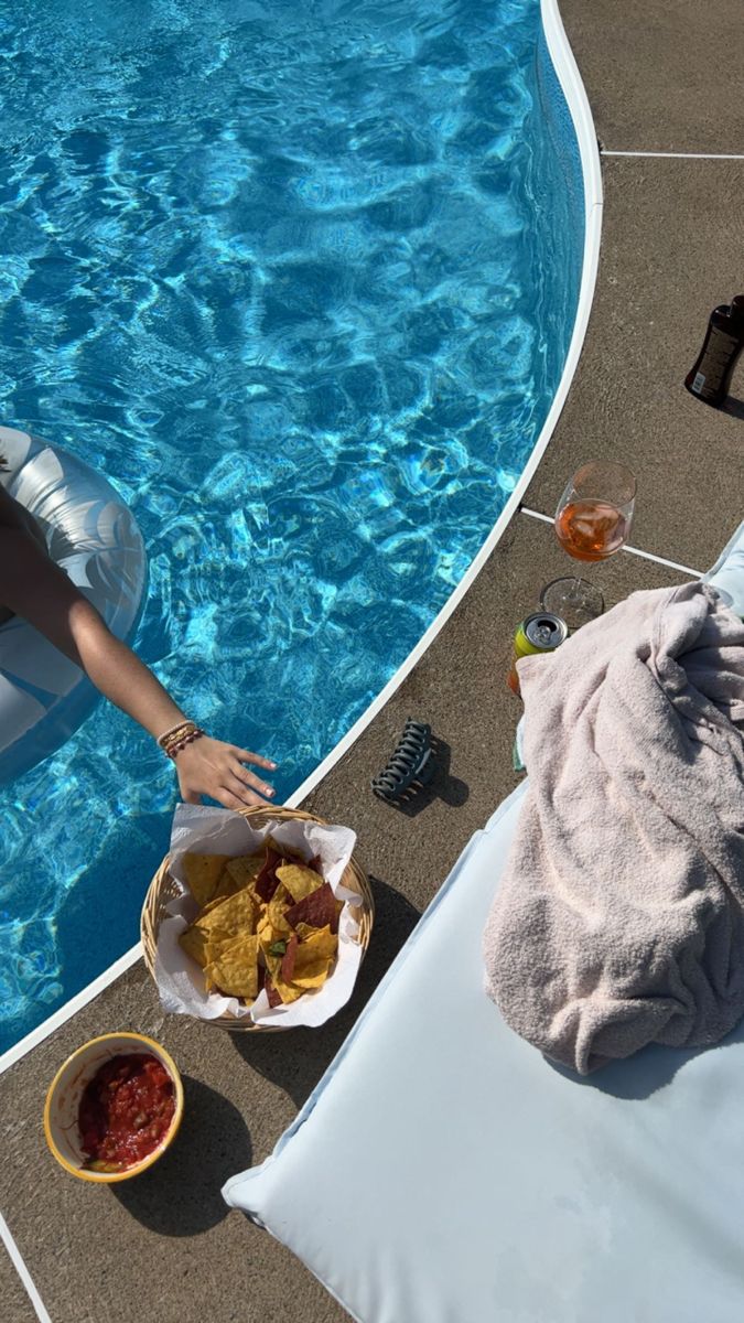 a woman sitting in front of a swimming pool holding a basket of food and chips