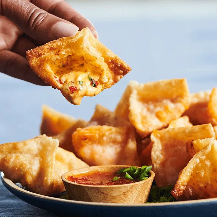 a hand holding a piece of deep fried food over a plate with dipping sauces