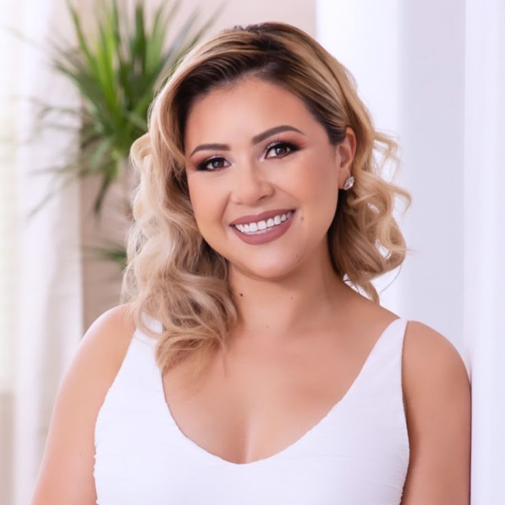 a woman with blonde hair wearing a white tank top and smiling at the camera while standing in front of a potted plant