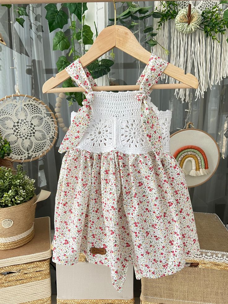 a white dress with red flowers is hanging on a wooden hanger in front of some potted plants