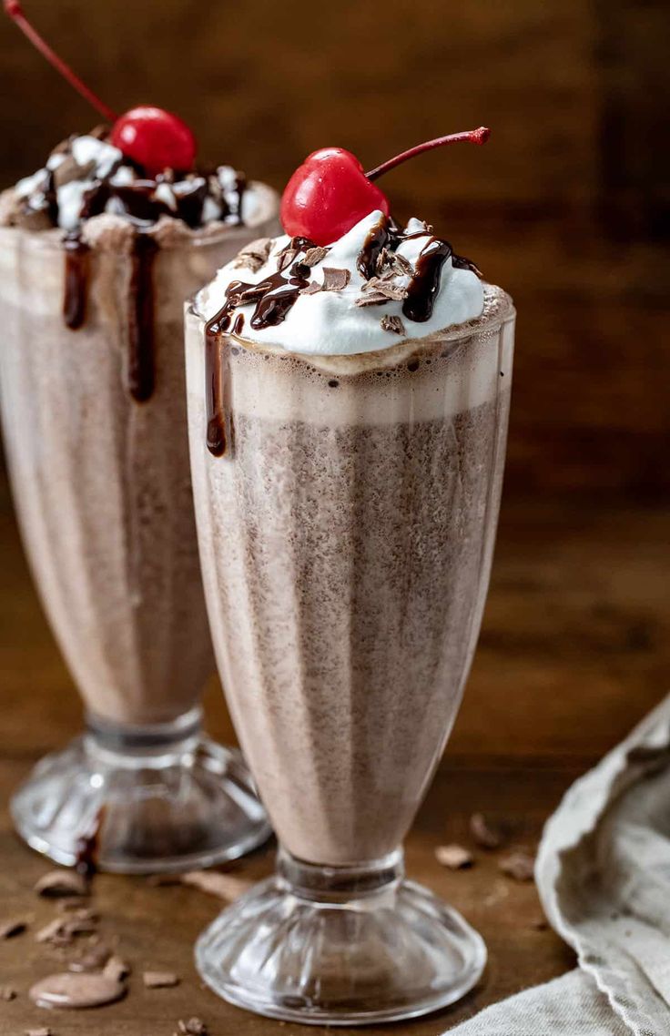 two glasses filled with chocolate milkshakes and topped with whipped cream and cherries