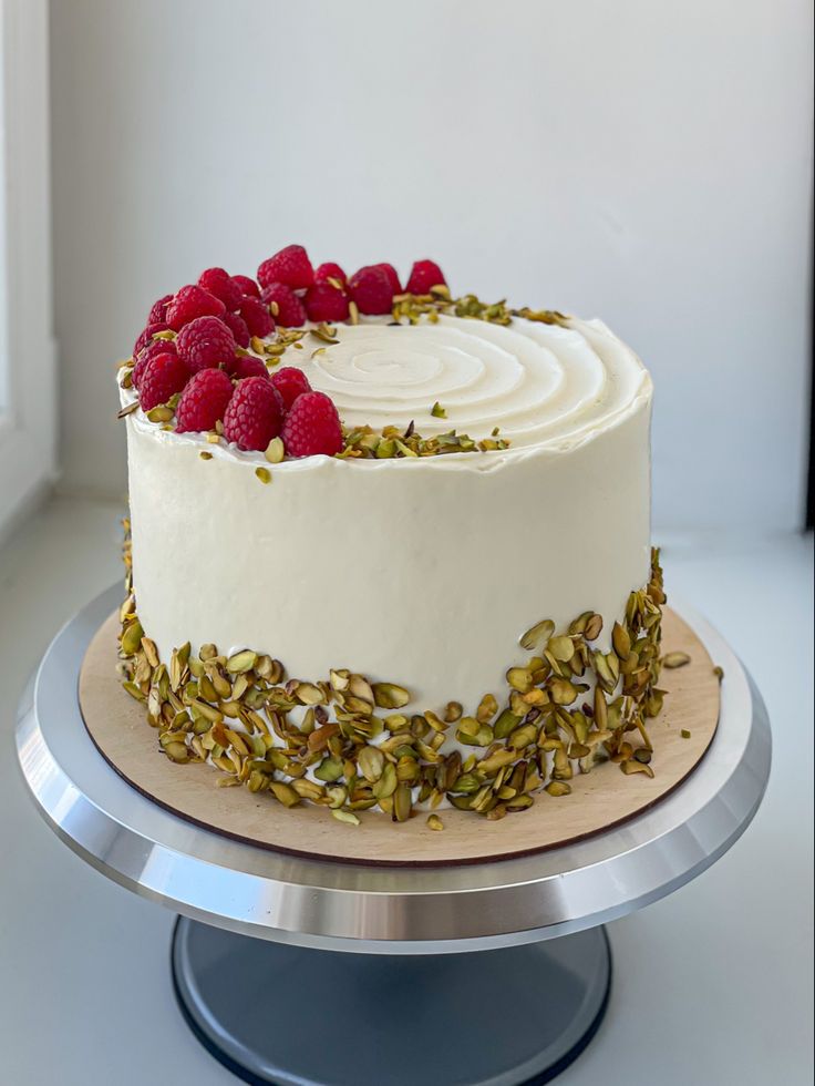 a white cake with raspberries and pistachios on top sits on a silver platter