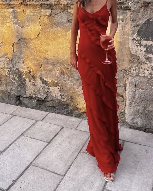 a woman in a red dress holding a wine glass and standing next to a stone wall