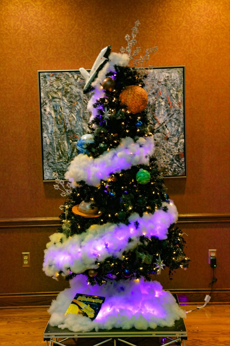 a decorated christmas tree with purple lights and snow on the top, in front of a painting