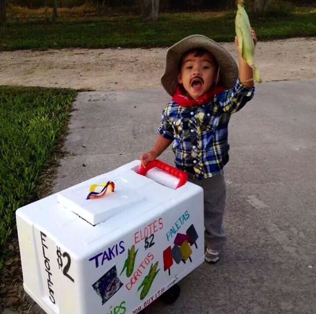 a young boy holding up a banana next to an ice chest on the sidewalk with grass and trees in the background