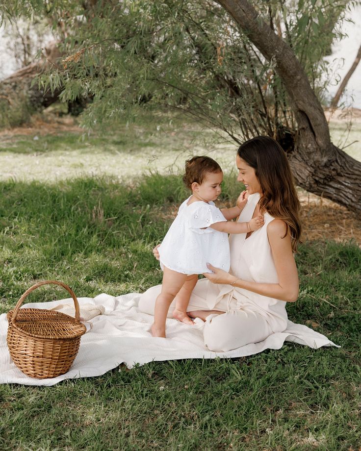 a woman holding a baby while sitting on a blanket in the grass near a tree