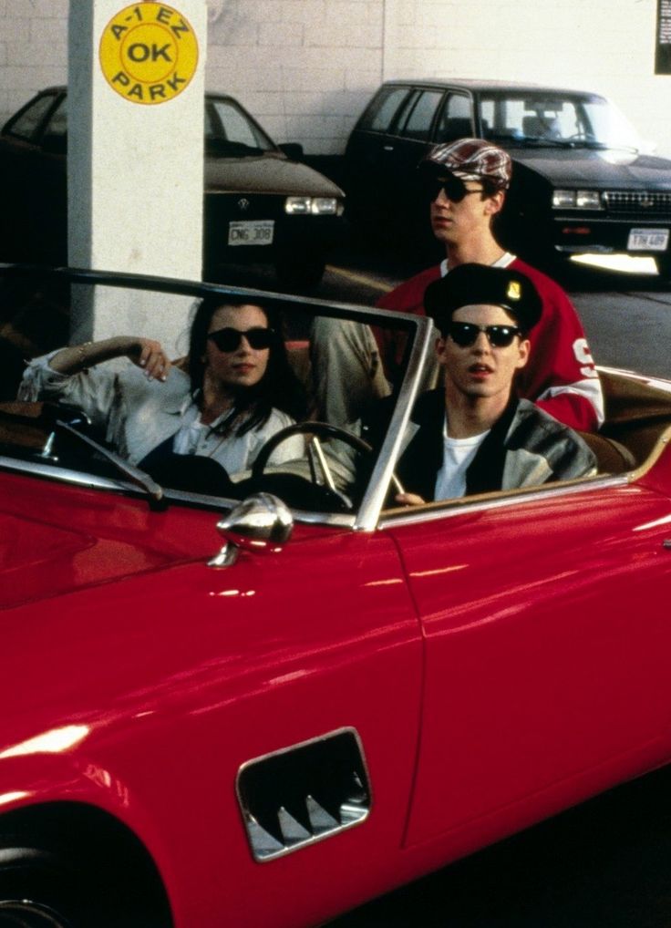 three men sitting in a red convertible car with the driver looking at the camera while another man stands next to them