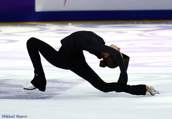 a male figure skating on an ice rink during a game in the winter olympics, with one leg bent over his head