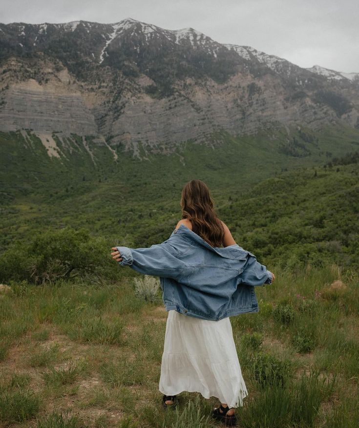 a woman is standing in the grass with her arms spread out and mountains behind her