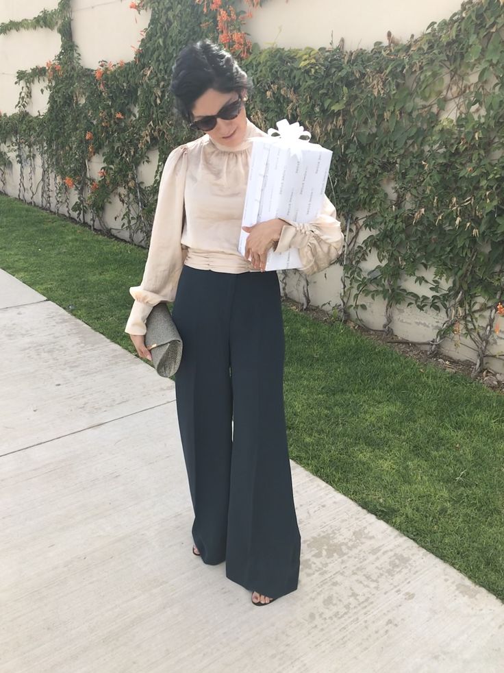 a woman is standing on the sidewalk and holding some papers in her hand while wearing black wide legged pants