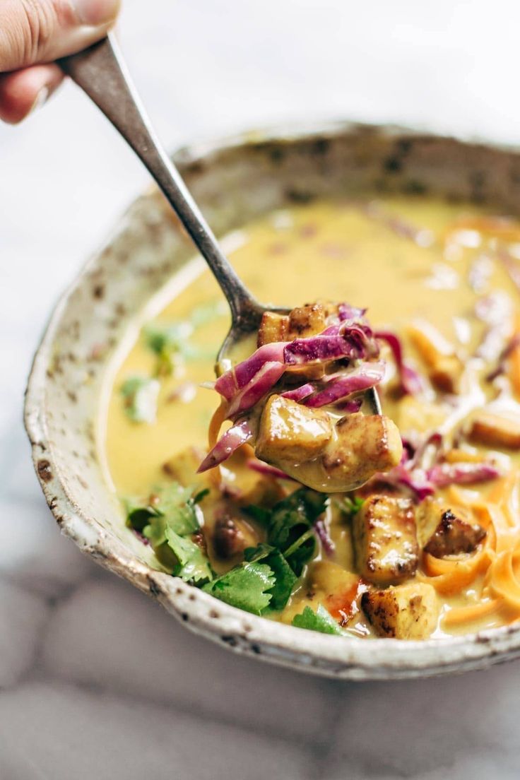 a hand holding a spoon full of soup with tofu and noodles in it on a marble table