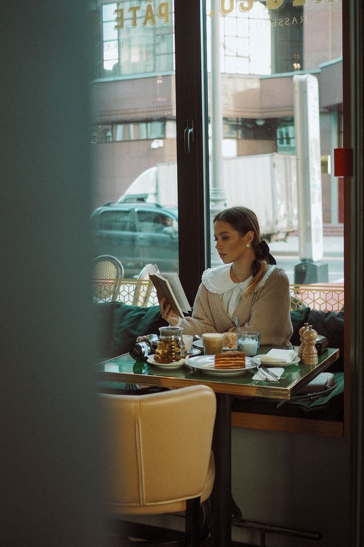 a woman sitting at a table in front of a window reading a book and eating food