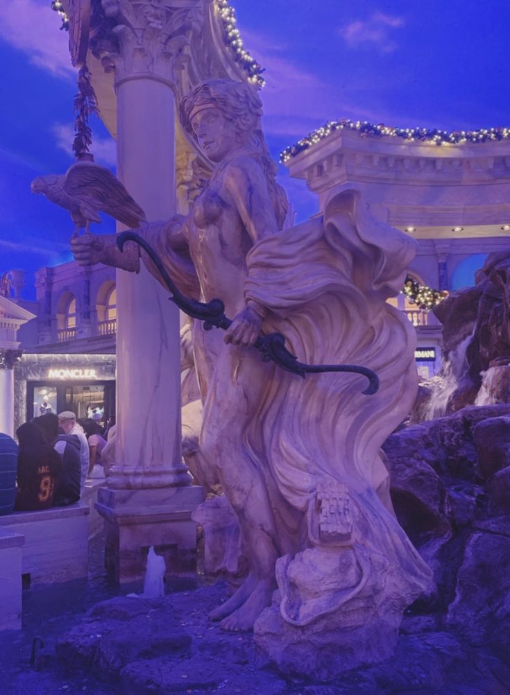 a statue in the middle of a building with lights on it's sides and people walking around