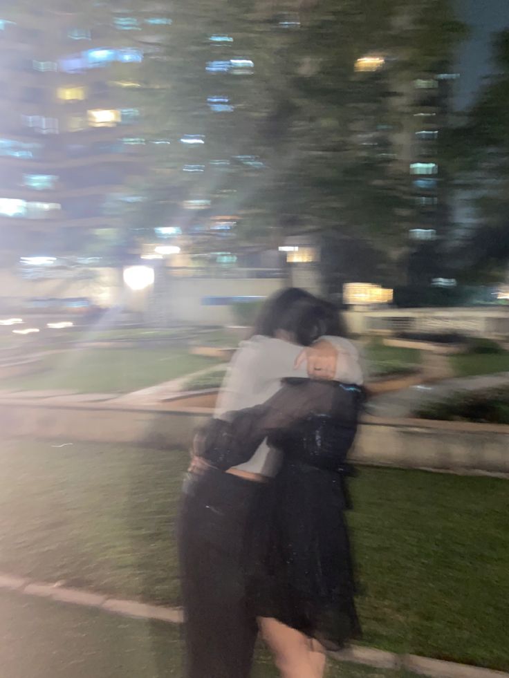 two people hugging each other in front of a cityscape at night with buildings and lights behind them