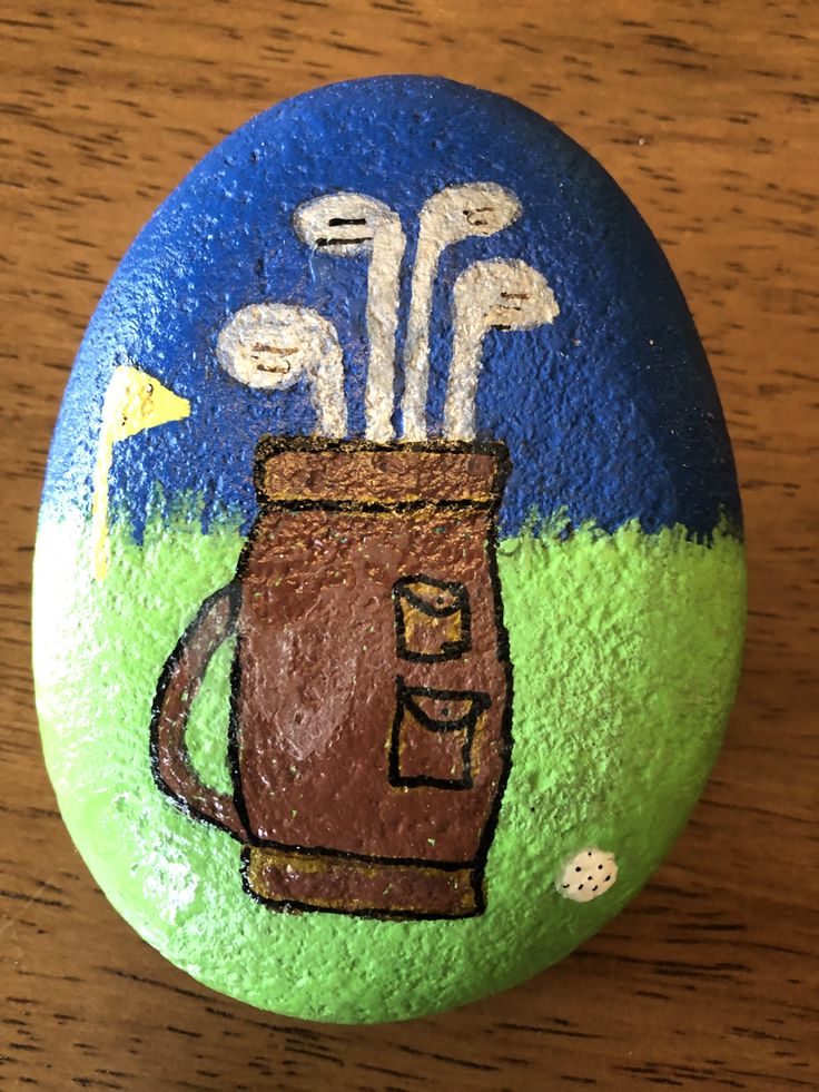 a painted rock with an image of golf clubs in a cup on it, sitting on a wooden table