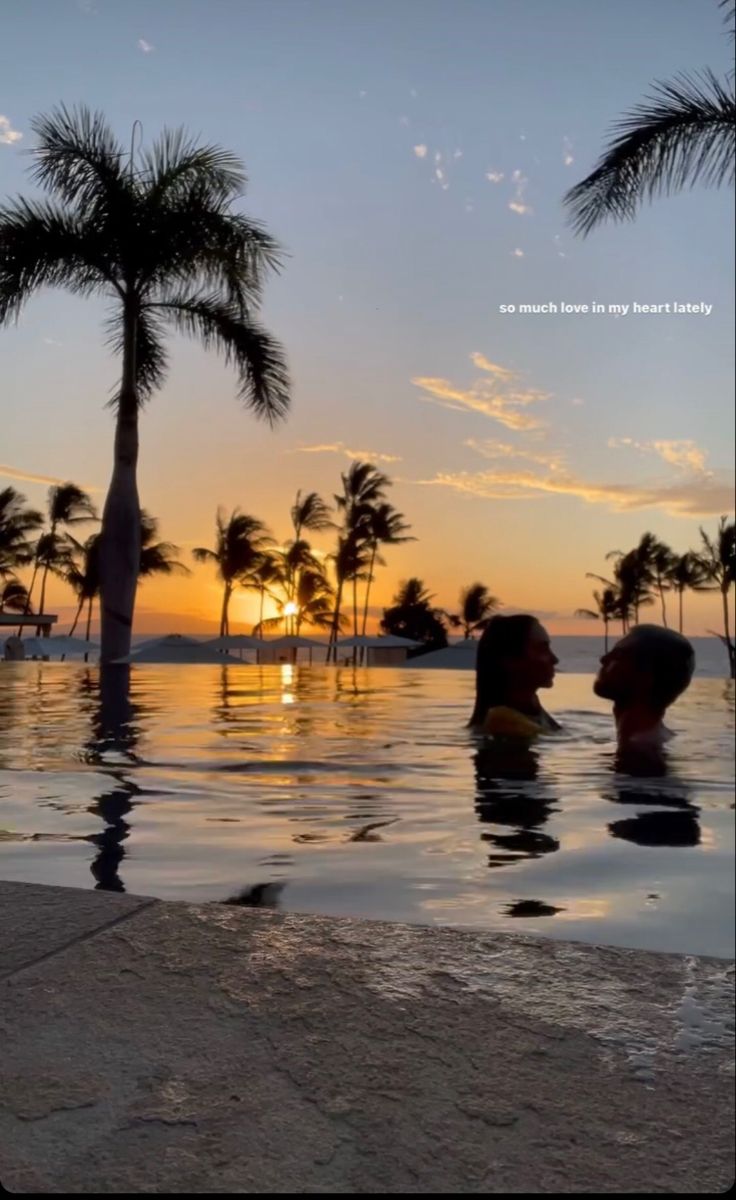 two people are sitting in the pool at sunset with palm trees and water reflecting them